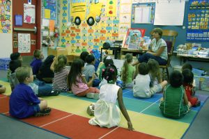 A teach reads a book aloud to a group of children sitting on a carpet.