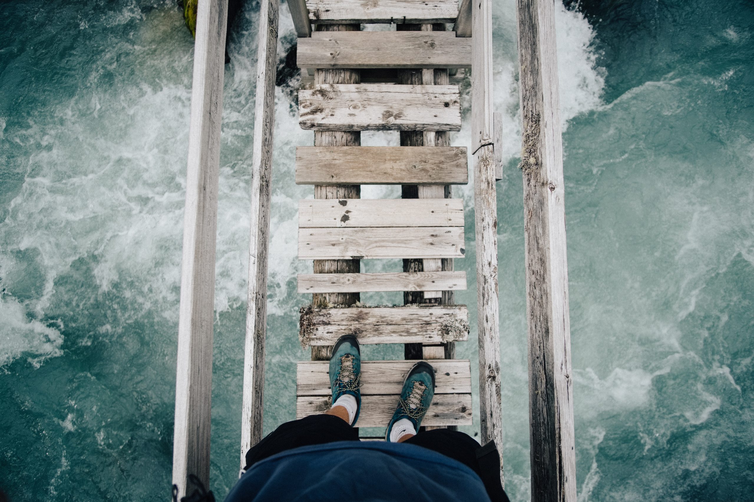 Person standing on unstable wooden bridge over rough waters