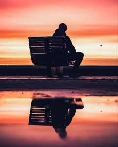 A person is sitting outside watching a sunset, and the image is reflected in a lake or pond.