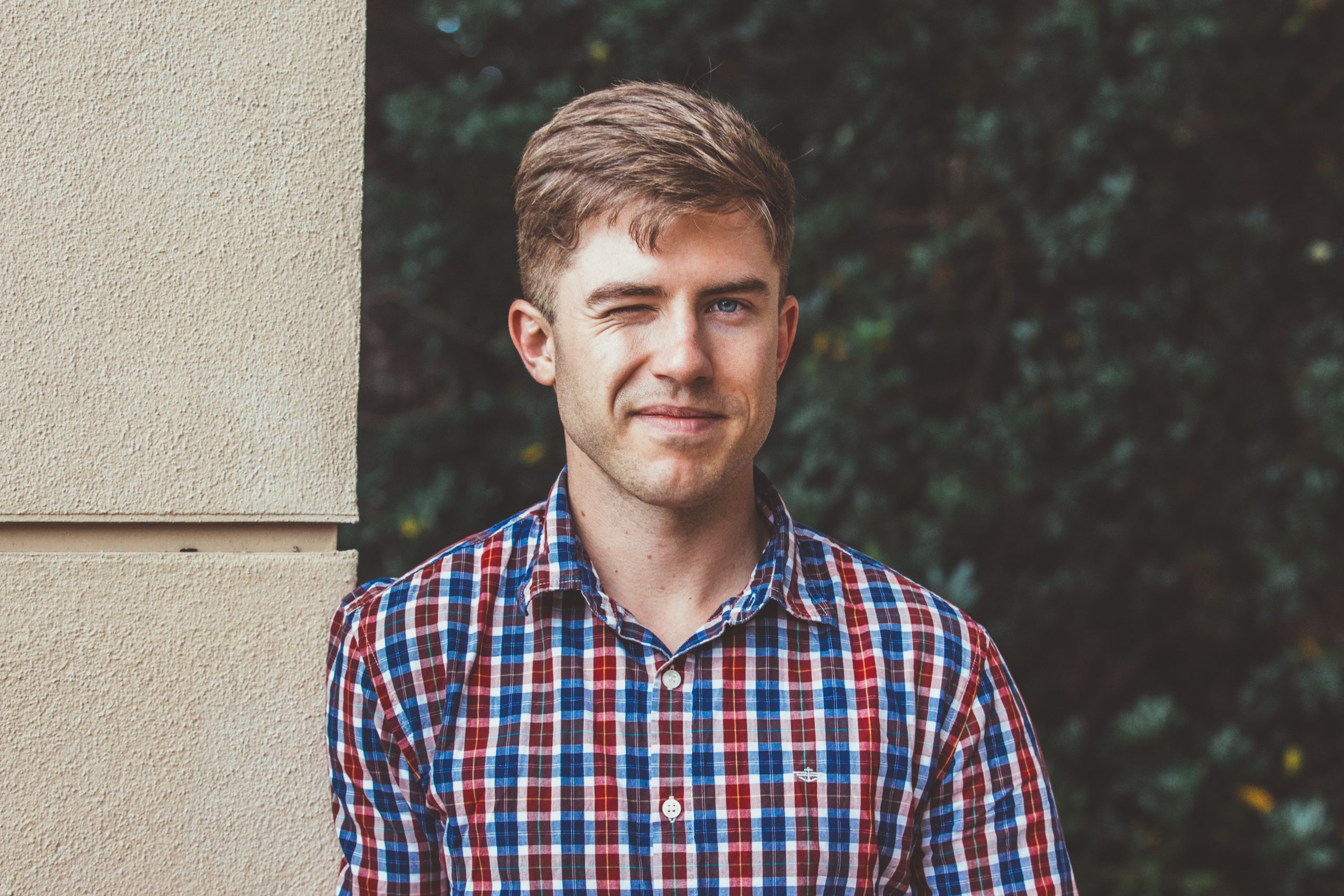 Man in a plaid shirt leaning against an outdoor post. He is winking.