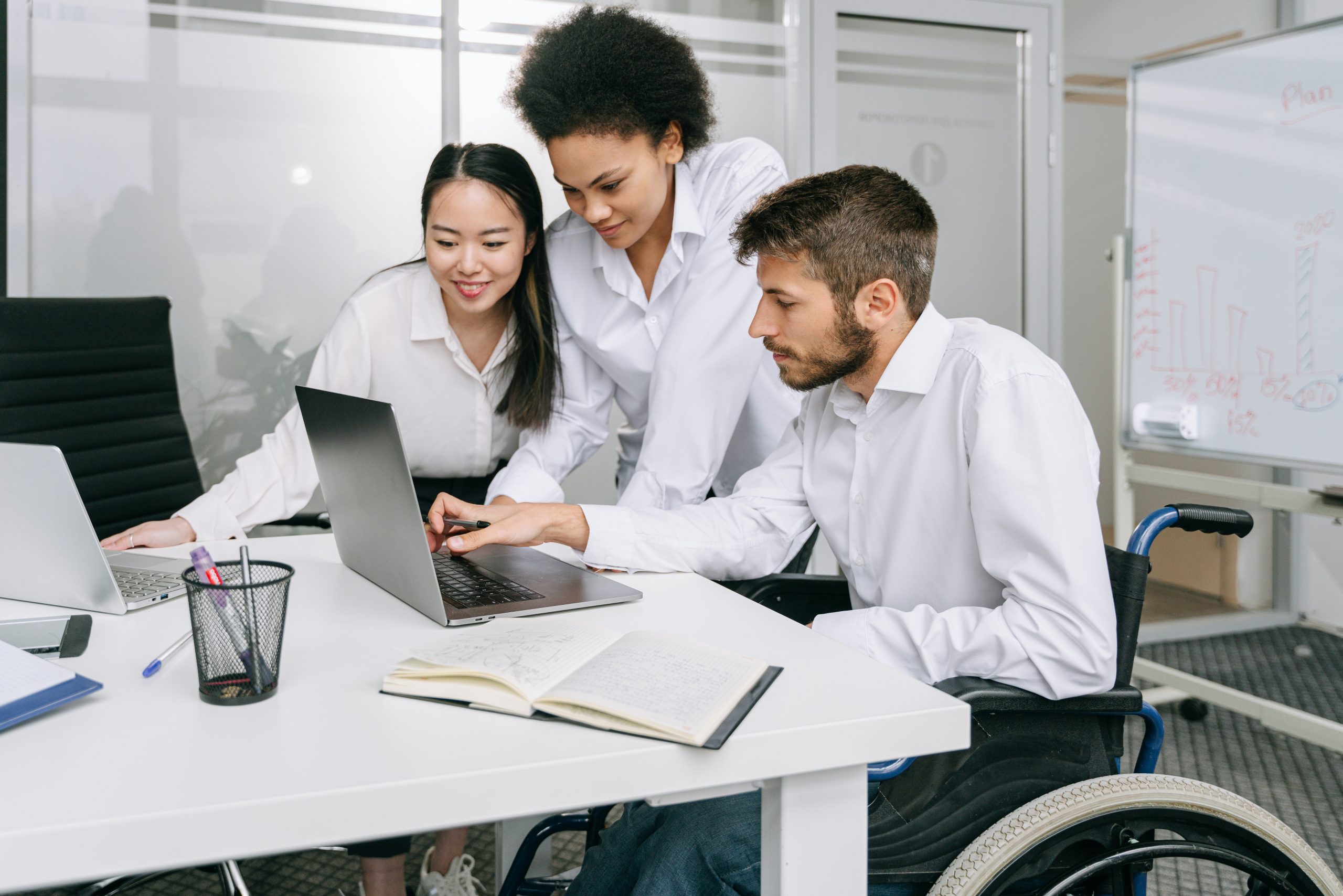 A white man with a wheelchair, Asian woman, and Black woman are all smiling and looking at a computer screen.