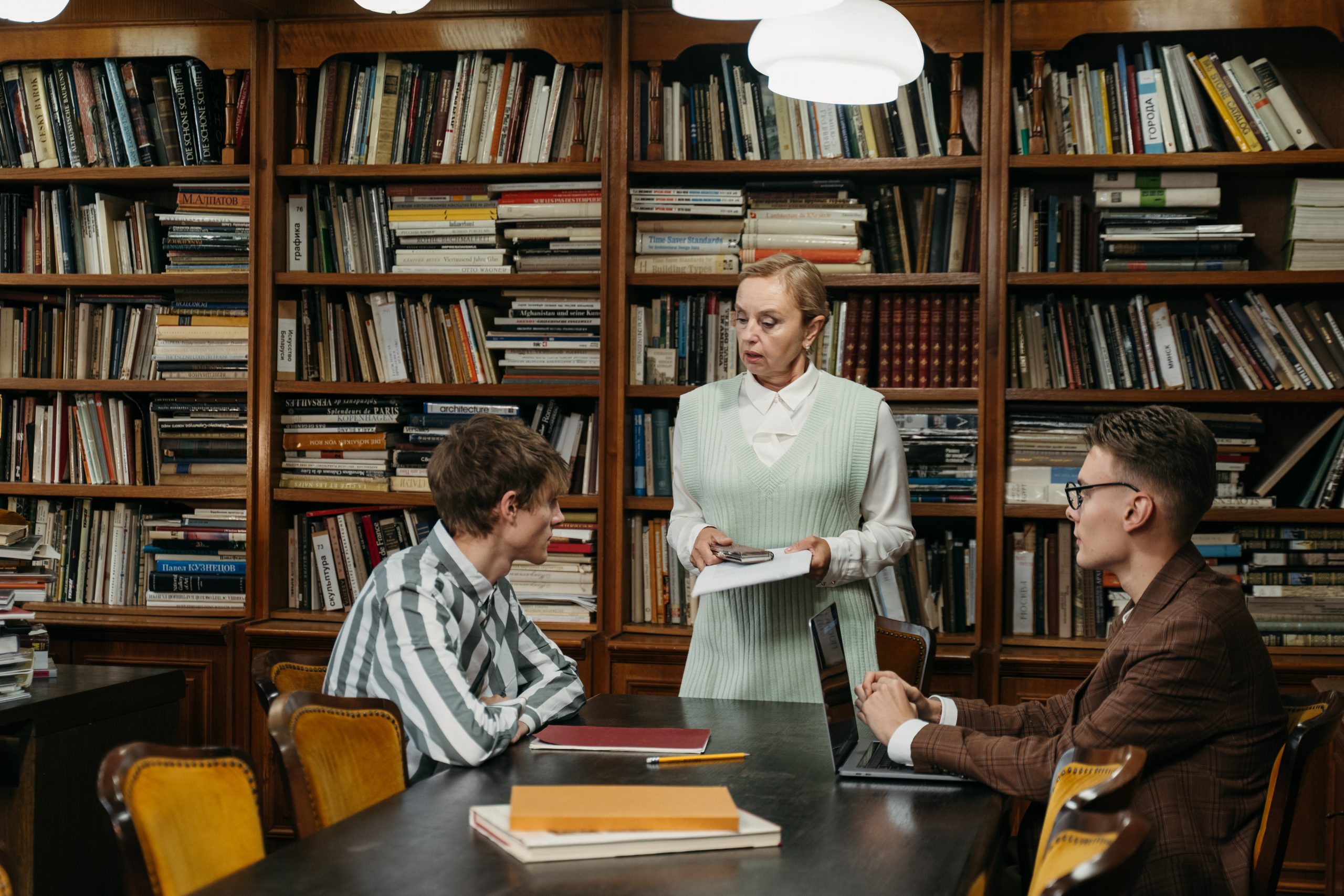 Two met sit at a table in the library. A older female librarian is standing beside the table speaking to them. Books line the walls in the background.