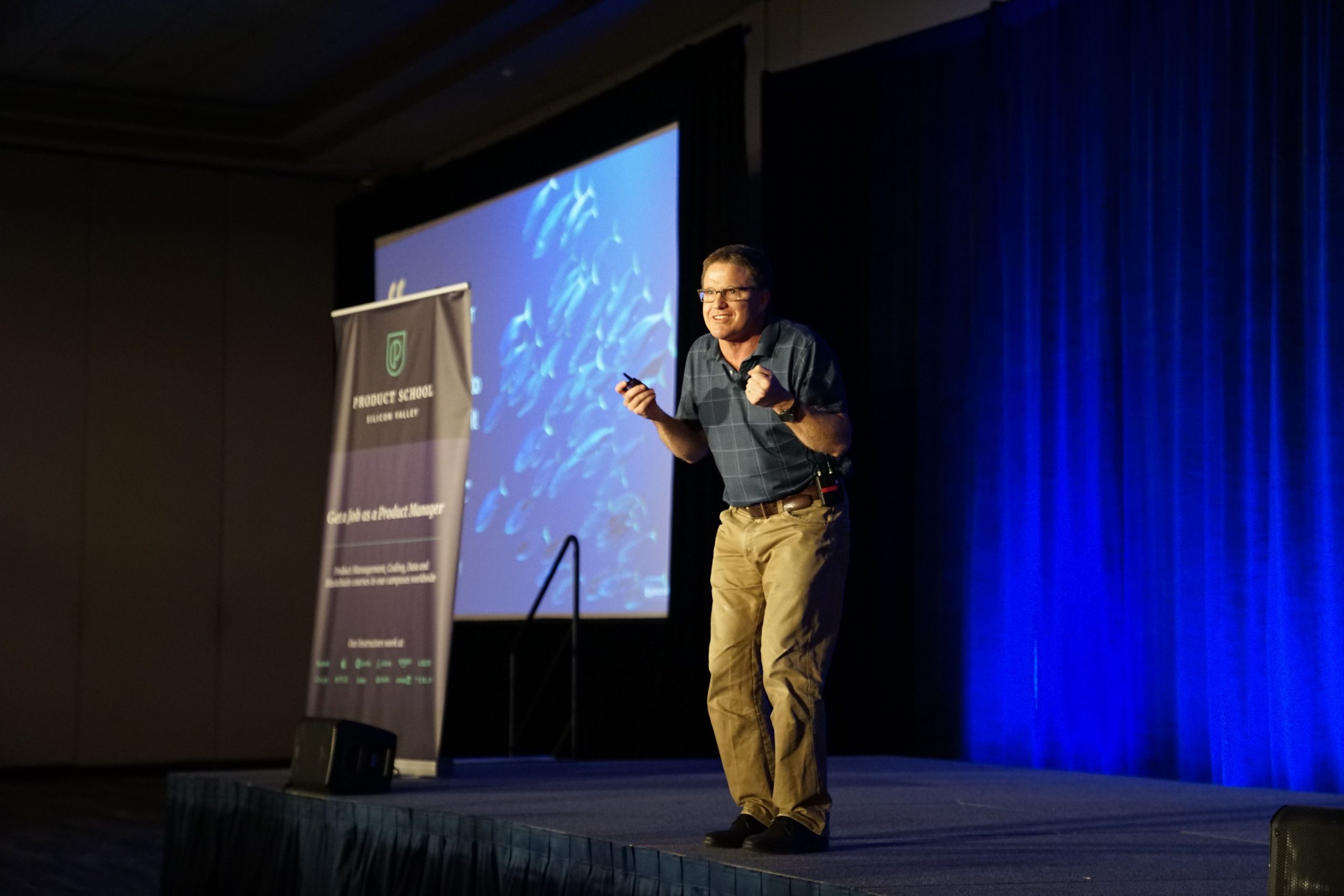 Man on stage with a powerpoint clicker in one hand, standing away from the podium with his legs bent a little, as if almost ready to walk.