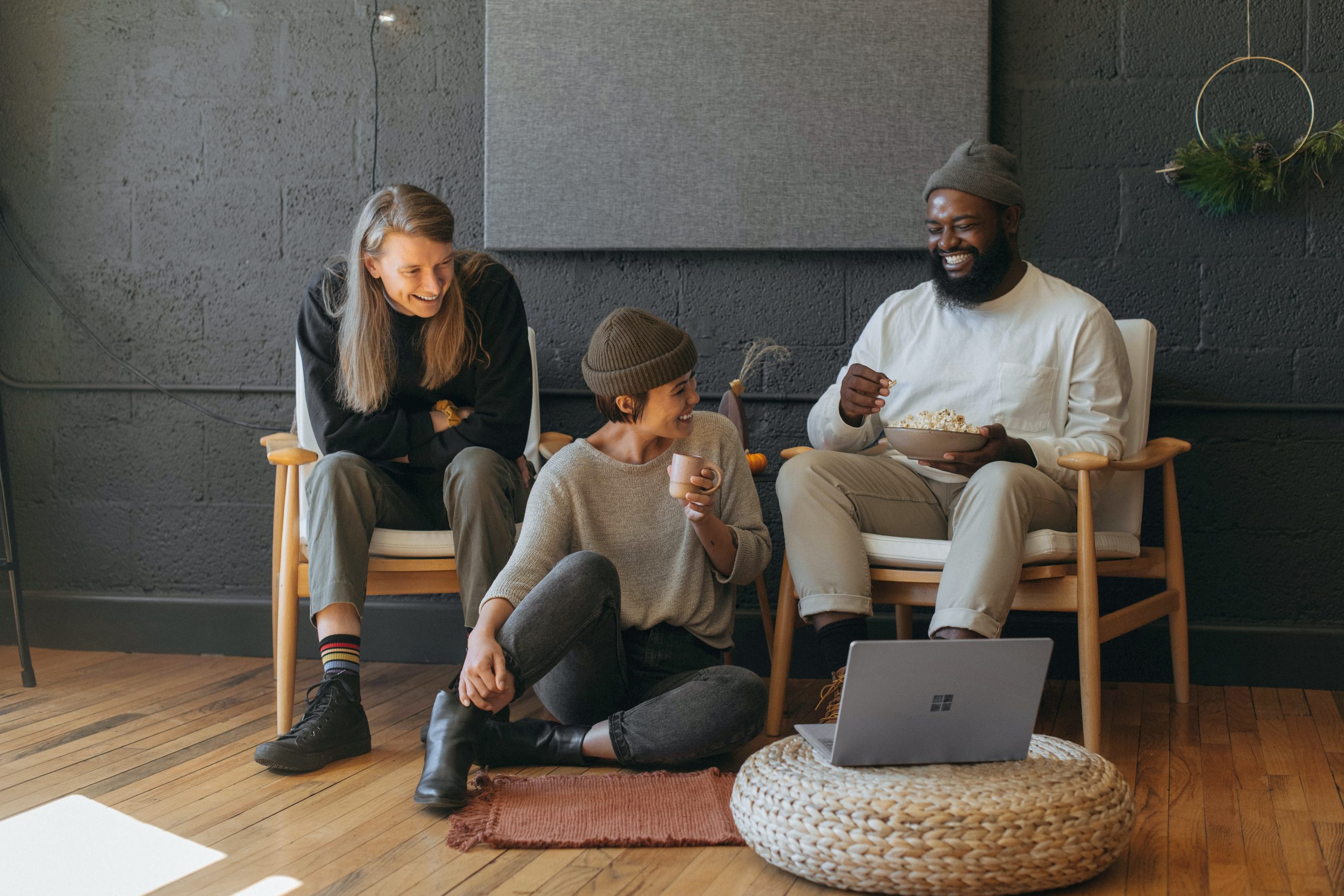 Three friends sitting in chairs and on the floor eating, drinking coffee, laughing, and looking at a laptop.