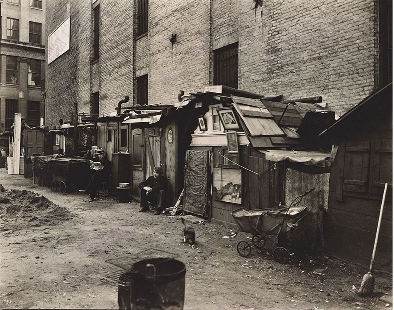A black and white photograph captures a street scene in Lower Manhattan dominated by makeshift shelters or shanties built against a brick building on the left. The ground is dirt, suggesting a temporary or impoverished settlement. Several figures are visible; two men are seated outside the shanties, one appearing to be reading. A small dog walks in the foreground. To the right, a baby carriage and a broom are visible near another shelter.