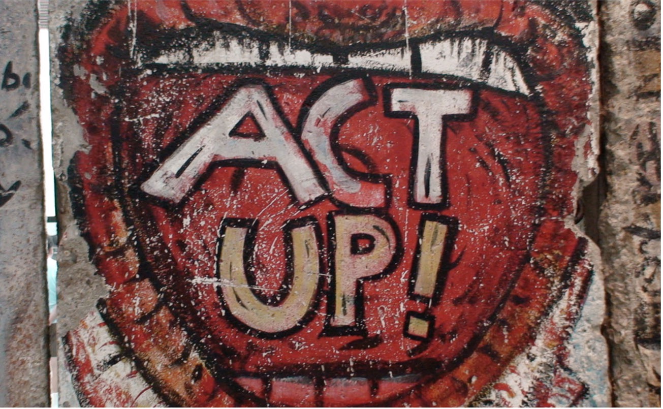 Close-up of red and white graffiti on a wall that says "ACT UP!" within a stylized mouth shape.