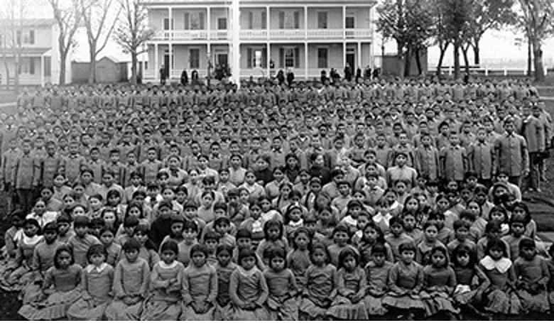 Photograph of a large number native children dressed in western style school uniforms posed in front of a white columned building.