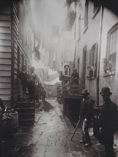 Jacob Riis's photograph of Bandit’s Roost an alley way in New York city. It depicts men lined up on either side of the alley with children and woman leaning out of a window.
