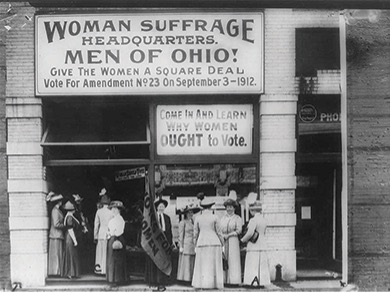 Photograph of women standing outside of a building with a sign that says "Women Suffrage / Headquarters / Men of Ohio! / Give the Women a Square Deal / Vote for Amendment No. 23 on September 3 - 1912" Beneath that sign in another that states "Come in and learn / why women / ought to vote."