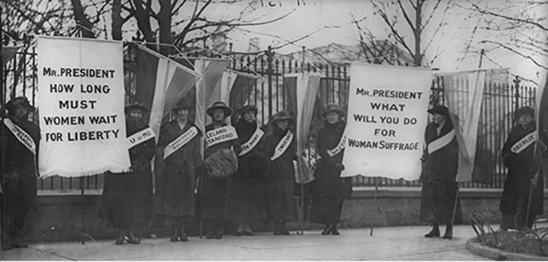 Photograph of Alice Paul and other members of the Women's Party wearing sashes with slogans on them. On the left and the right women hold two banners. One reads "Mr. President how long must women wait for Liberty" and the other reads "Mr. President what will you do for Women Suffrage."