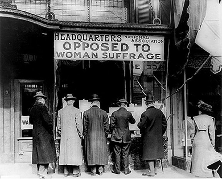 Photograph of men standing outside the anti-suffrage headquarters peering into a winder. Above them is a sign that reads "Headquarters National Association / Opposed To / Woman Suffrage."