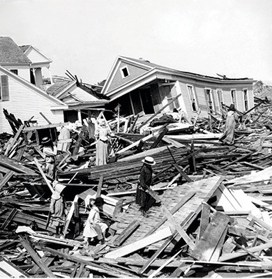 Photograph of a house destroyed by a hurricane. People climb over the rubble of wood and roof matter.