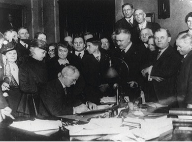 A black and white photo of Governor James Goodrich seated at at table signing the bill to prohibit alcohol in the state. Surrounding him are men and women who supported the bill.