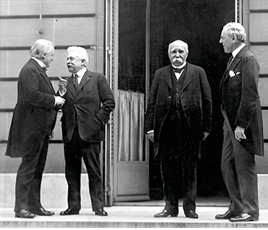 The image shows a black and white photograph of the Paris Peace Conference. The following public figures are identified: Vittorio Emanuele Orlando and David Lloyd George. The image captures four men in suits standing outside a building, engaged in conversation.