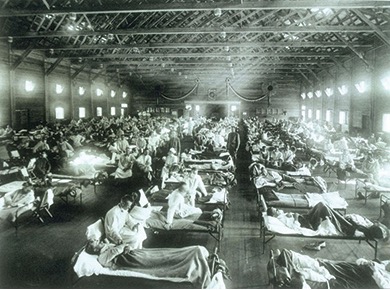 A black and white photo showing a temporary hospital ward with rows of beds. Many people are lying in the beds, and nurses are attending to them. This depicts a scene during the Spanish flu pandemic.
