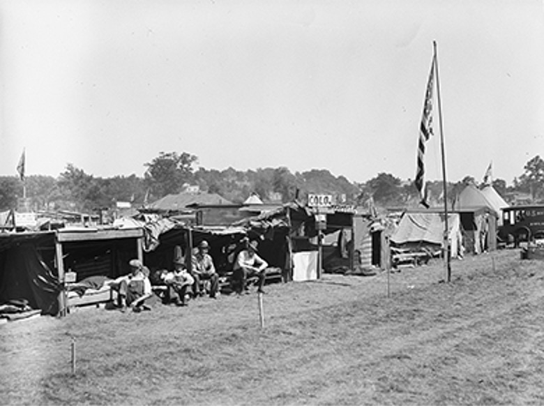 A black and white photograph capturing a wide view of a Bonus Army encampment. Numerous makeshift shacks and tents, constructed from various scavenged materials like wood, tarps, and corrugated metal, stretch across a grassy field under a bright sky. Several men are visible sitting or standing outside their shelters. A tall flagpole with a large American flag stands prominently in the foreground on the right. In the distance, trees and other structures can be seen. The scene conveys the temporary and improvised nature of the camp.