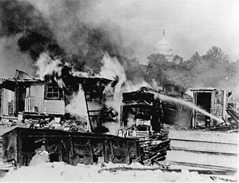 A black and white photograph shows several makeshift structures on fire, sending thick plumes of smoke into the sky. A stream of water from a hose is directed towards one of the burning buildings. In the background, partially obscured by trees, the dome of the U.S. Capitol Building is visible, suggesting the location of the Anacostia Flats camp of the Bonus Army. The scene depicts the destruction of the encampment.