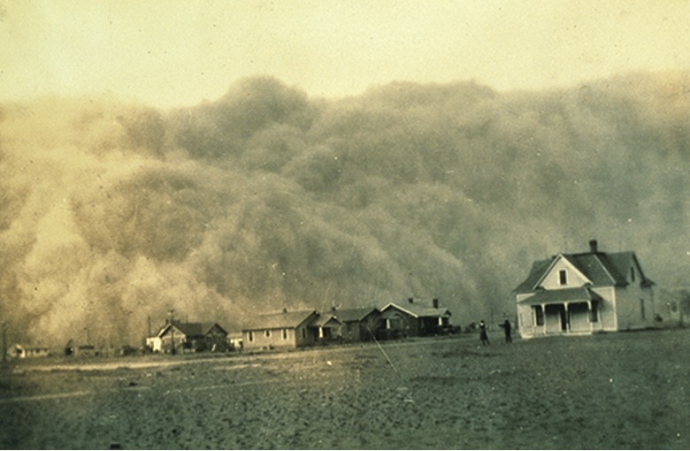 A sepia-toned photograph captures a massive dust storm engulfing a rural landscape during the Dust Bowl era. A colossal cloud of dust, appearing like a towering wall, rolls across the horizon, obscuring much of the sky. In the foreground, a few scattered houses and buildings stand on a barren, flat plain, dwarfed by the immense dust cloud. Two figures are visible near a house on the right, seemingly observing the approaching storm. The image conveys the overwhelming power and destructive nature of a dust storm.