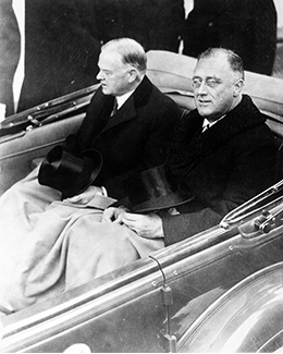 A black and white photograph of two men, identified as Franklin D. Roosevelt and Herbert Hoover, sitting side-by-side in the back seat of an open-top car. Both men are dressed in dark suits and coats, and each holds a top hat on their lap. Franklin D. Roosevelt, on the right, appears to be looking towards the viewer with a slight smile, while Herbert Hoover, on the left, is looking forward with a more serious expression. The image likely captures them together during a formal event or transition of power.