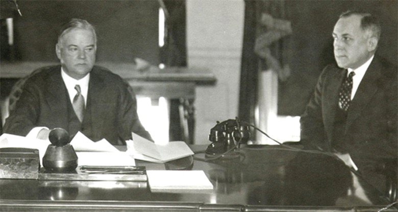 A black and white photograph shows two men, including Herbert Hoover, seated at a large, dark wooden desk in what appears to be an office. Herbert Hoover, on the left, is seated facing forward, looking slightly off to the left, with papers and an inkwell on the desk in front of him. The man on the right, who is not identified, is looking intently towards Herbert Hoover. Both men are dressed in suits and ties. The background shows a window with drapes and what appears to be part of a column or architectural detail. The scene suggests a formal meeting or work session.