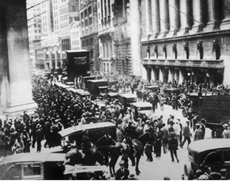 A black and white photograph capturing a bustling street scene, likely outside the New York Stock Exchange during the Wall Street Crash of 1929. A massive crowd of people, predominantly men in suits and hats, fills the street and sidewalks. Several vintage cars and horse-drawn carriages are visible amidst the crowd. On the right, the grand facade of a large, classical building with columns and statues can be seen, presumed to be the NYSE. The scene conveys a sense of urgency and significant public presence during a historical financial event.