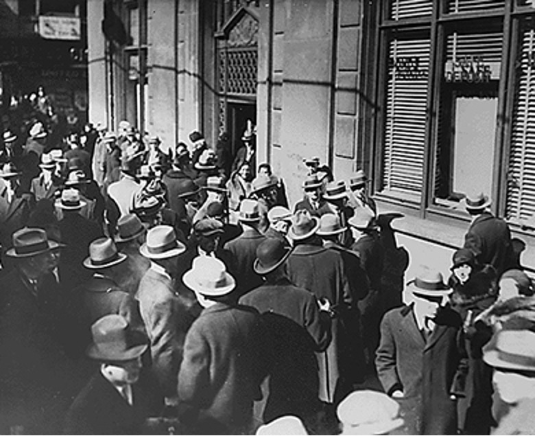 A black and white photograph shows a dense crowd of men, most wearing hats and suits or coats, gathered on a city sidewalk outside a building. Many are looking intently towards a window or entrance, while others are in conversation. The building has large windows with horizontal blinds partially drawn, and what appears to be a doorway to the left. The lighting suggests it is daytime, with strong shadows on the building's facade. The atmosphere seems to be one of anticipation or concern.