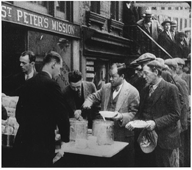 A black and white photograph shows a group of men gathered outside "St. Peter's Mission," where two individuals are serving food from large containers on a table. A line of men, some wearing hats, are waiting with bowls or plates to receive food. The building behind them appears to be a brick structure, and the overall scene suggests a soup kitchen or charitable food distribution during a time of hardship, such as the Great Depression.