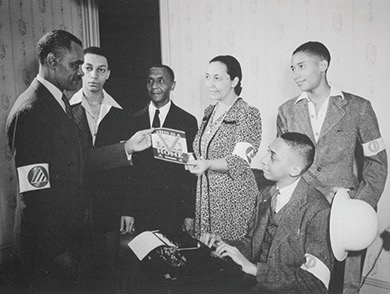 A black and white photo of a group of six individuals gathered around a table with a typewriter. One man on the left is holding up a pamphlet or card that appears to say "DOUBLE V IS FOR VICTORY AT HOME AND ABROAD". Several people in the group are wearing armbands with a symbol. Based on visual evidence and historical context often associated with this image, the individuals appear to be Black and involved in the Double V campaign.