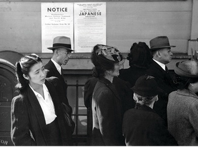 A black and white photo showing a group of Japanese Americans standing in a line on a sidewalk as part of the Japanese American internment process. Signs in the background have notices regarding "Western Defense Command and Fourth Army" and "Instructions to all persons of JAPANESE ANCESTRY."