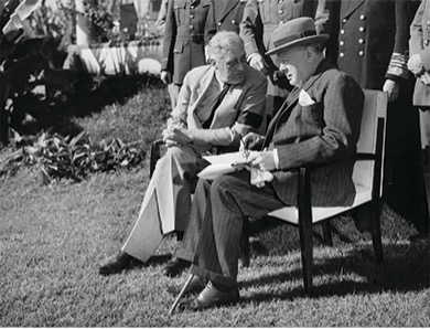 A black and white photo of Winston Churchill and Franklin D. Roosevelt seated outdoors. They are both wearing coats and hats and appear to be in discussion. Other military figures are standing behind them.