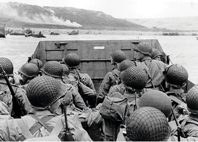 A black and white photo looking over the shoulders of soldiers in helmets, packed into a landing craft. They are approaching a beach where other landing crafts and vehicles are visible, depicting a scene from the D-Day landings.