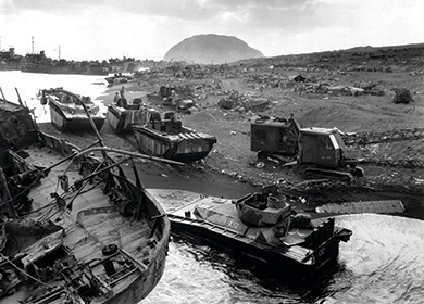 A black and white photo showing a beach littered with damaged landing vehicles and equipment. The water's edge shows more vehicles, and a large hill is visible in the background. This depicts a scene from the Battle of Iwo Jima.