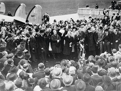 A black and white photo of Neville Chamberlain stepping off an airplane and being greeted by a large crowd of people.