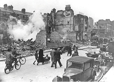A black and white photo showing a street scene with damaged and collapsed buildings, debris, and smoke, likely depicting the aftermath of an air raid during the Blitz. People are walking on the street, including a family with a baby carriage.