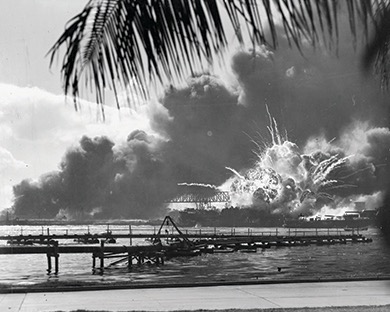A black and white photo showing an explosion on the water with smoke billowing in the background. Structures along the shore and palm fronds are visible in the foreground, depicting the attack on Pearl Harbor.