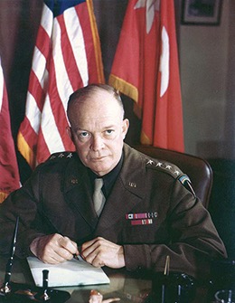 A color portrait of Dwight D. Eisenhower in uniform, seated at a desk with an American flag and another flag behind him. He is looking directly at the viewer and holding a pen over a paper.