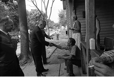 A black and white photo of Lyndon B. Johnson shaking hands with a man seated on a porch. Other people are standing nearby. This scene appears to depict Johnson interacting with residents as part of his Great Society initiatives.