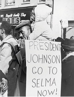 A black and white photo of a black man carrying a child on his shoulders during a demonstration. The man is holding a sign that reads "PRESIDENT JOHNSON GO TO SELMA NOW!".