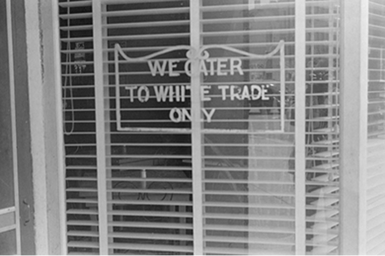 A black and white photo showing a sign in a window with blinds behind it. The sign reads "WE CATER TO WHITE TRADE ONLY."