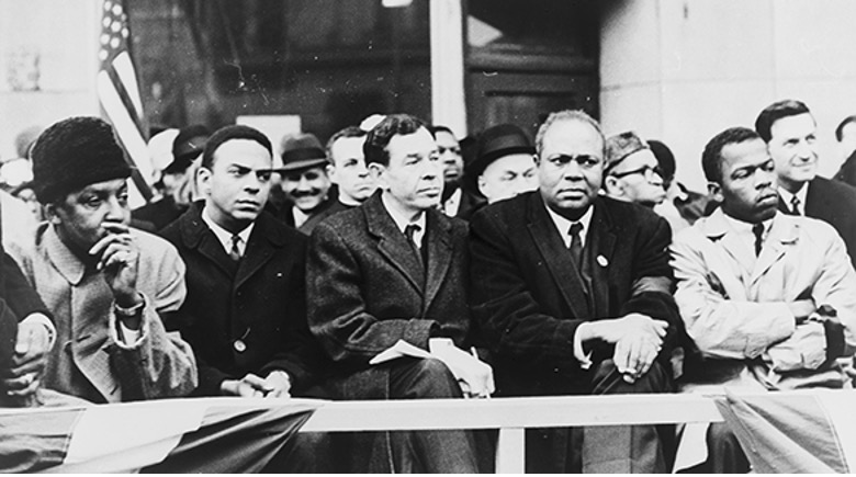 A black and white photo of a group of men, Civil rights activists Bayard Rustin, Andrew Young, Rep. William Fitts Ryan, James Farmer, and John Lewis (l to r), sitting in the stands.