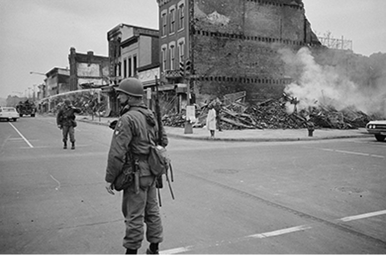 A black and white photo showing a street scene with damaged buildings and debris. Smoke is rising from some areas, and people are visible on the street. This depicts the aftermath of civil unrest, related to the King assassination riots based on historical context.