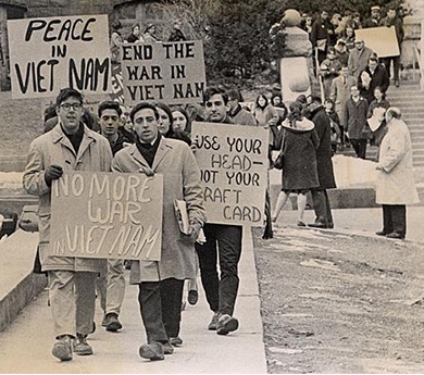 A black and white photo of a group of people, appearing to be mostly young adults, marching outdoors. They are holding signs protesting the Vietnam War. Signs read "PEACE IN VIET NAM", "END THE WAR IN VIET NAM", "NO MORE WAR IN VIET NAM", and "USE YOUR HEAD- NOT YOUR DRAFT CARD". They are walking on a sidewalk with buildings and steps in the background.