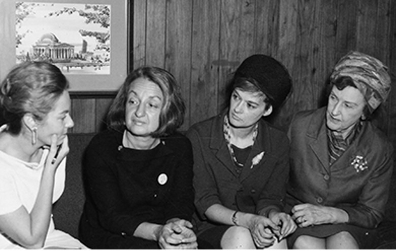 Four women of NOW, including Betty Friedan, sit together. A framed picture of a building is on the wall behind them.