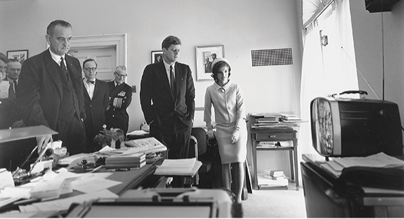 A black and white photo of John F. Kennedy, Jacqueline Kennedy, and Lyndon B. Johnson, along with other people, looking at a television set in a room. There is a desk in the foreground.
