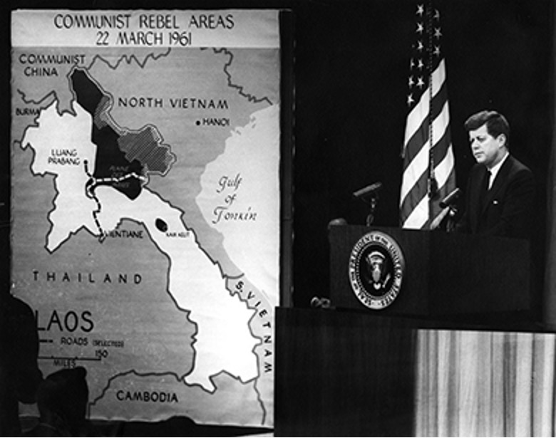 A black and white photo of John F. Kennedy standing at a podium with an American flag behind him, speaking in front of a large map showing "COMMUNIST REBEL AREAS" in Laos, North Vietnam, Thailand, and Cambodia.
