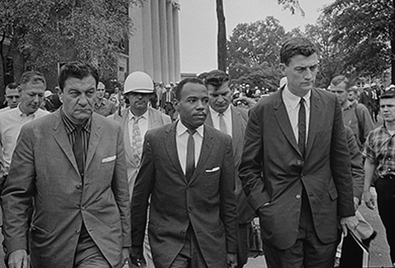 A black and white photo of several men walking outdoors. In the center is James Meredith, surrounded by other men, some of whom appear to be accompanying him, and others who appear to be onlookers or security as he integrated the University of Mississippi.