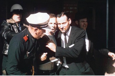 A color photo showing Jack Ruby, restrained by a police officer as he lunges forward toward Lee Harvey Oswald who is also being held by a man in a suit. This captures the moment Lee Harvey Oswald was shot.