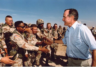 A color photo of George H.W. Bush in shirt sleeves,, standing with a group of soldiers in desert camouflage uniforms shaking the hand of one during the Gulf War.