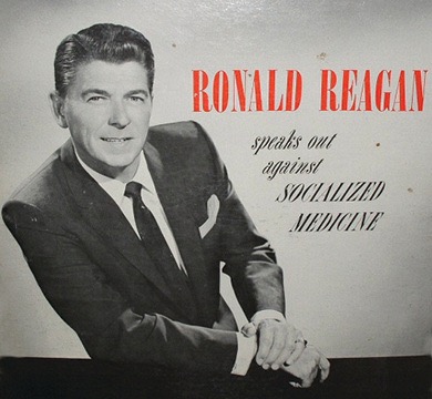 A black and white photo of Ronald Reagan standing at a podium with microphones, speaking to an audience. A large sign behind him reads "SOCIALIZED MEDICINE.