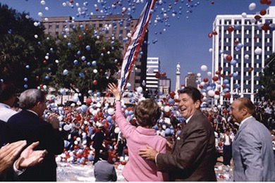 A color photo of Ronald Reagan and Nancy Reagan standing together outdoors. Ronald Reagan is wearing a suit and Nancy Reagan is wearing a dress and holding a bouquet of flowers. They are both smiling and waving to a crowd in the background.